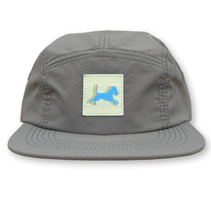 May include: Gray five-panel cap with a flat brim. A square patch on the front features a colorful dog silhouette. The dog is outlined in blue, with rainbow stripes. The cap is made of a smooth, durable material.