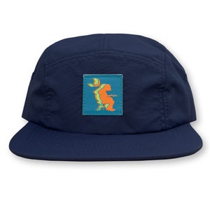 May include: A navy blue 5-panel cap with a patch featuring a stylized orange figure sitting on a stool with a crescent moon behind it. The patch has a blue background with green and yellow stripes.