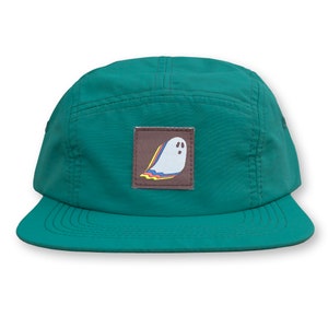May include: A teal green 5-panel cap with a brown patch featuring a white ghost with a rainbow design.
