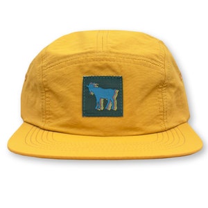 May include: A mustard yellow 5-panel cap with a dark green patch featuring a blue goat with rainbow stripes.