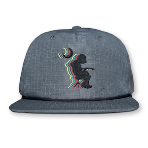 May include: A gray baseball cap with a black brim and a black embroidered design of a person sitting on a chair with a crescent moon behind them. The design is in a multi-colored, layered style.
