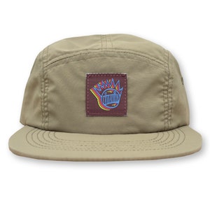 May include: A khaki colored 5-panel hat with a brown patch featuring a colorful cartoon face with a crown.