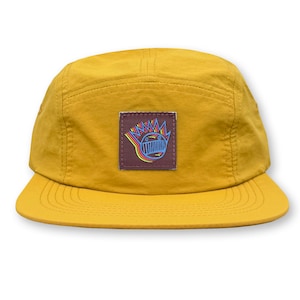 May include: A yellow 5-panel cap with a brown patch featuring a colorful graphic of a cartoon face with a crown of spikes.