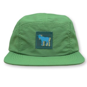 May include: A green five-panel cap with a flat brim. The cap features a square patch with a blue goat illustration with yellow and blue accents. The cap is made of a lightweight material.