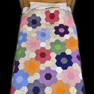 May include: A colourful, handmade quilt with a floral pattern covers a bed. The quilt features a repeating hexagon flower design in shades of pink, blue, orange, and purple. The bed has a wooden headboard and a white pillow. The quilt is made with a variety of fabrics.