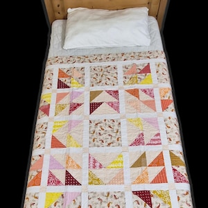 May include: A handmade quilt with a patchwork design featuring triangles in shades of orange, yellow, pink, and burgundy. The quilt is placed on a bed with a white pillow and a wooden headboard. The quilt's border has a repeating pattern of fish.