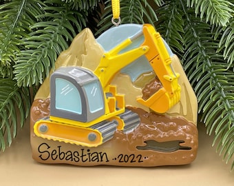 Pode incluir: Adorno de Natal de escavadora amarela com fundo de terra marrom e montanhas. A escavadora está cavando terra com seu balde. O adorno é personalizado com o nome "Sebastian" e o ano "2022".