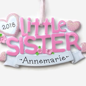 May include: Pink and white Christmas ornament with the words "Little Sister" and the year "2018" on a heart. The ornament also has a white banner with the text "...Annemarie..."