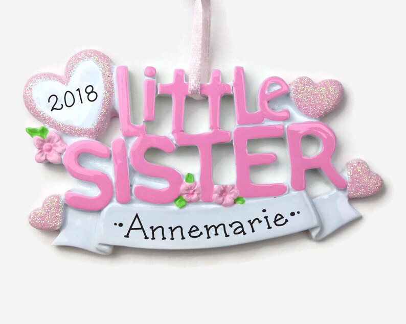 May include: Pink and white Christmas ornament with the words "Little Sister" and the year "2018" on a heart. The ornament also has a white banner with the text "...Annemarie..."