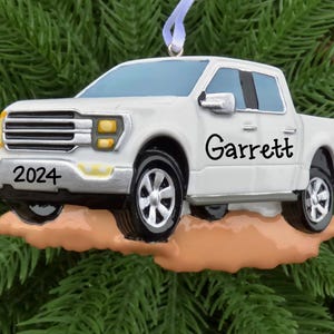 May include: White pickup truck Christmas ornament with the name "Garrett" and the year "2024" on the side. The truck is driving through a dirt patch.