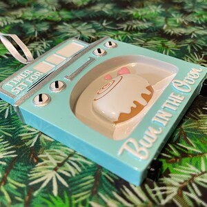 May include: A light blue ceramic ornament shaped like an oven with a cinnamon roll inside. The ornament has the text "Timer Set For Bun in the Oven" on the front.