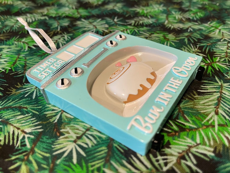 May include: A light blue ceramic ornament shaped like an oven with a cinnamon roll inside. The ornament has the text "Timer Set For Bun in the Oven" on the front.