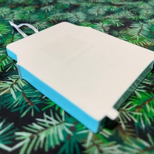 May include: A white blank ornament with a light blue border. The ornament has a string loop at the top for hanging.