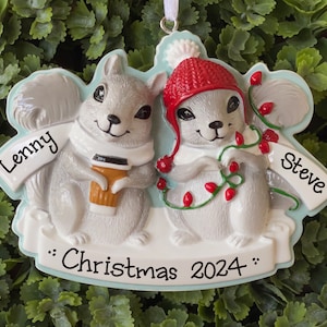 May include: A Christmas ornament featuring two smiling squirrels. One squirrel holds a coffee cup, the other is wrapped in Christmas lights. The ornament reads "Lenny", "Steve", and "Christmas 2024".