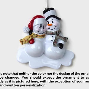 May include: A white ceramic Christmas ornament depicting a snowman couple. The female snowman is wearing a red hat and a plaid scarf, while the male snowman is wearing a black top hat and a gray scarf. Both snowmen are smiling and have black eyes and buttons. The ornament is on a white cloud-shaped base.