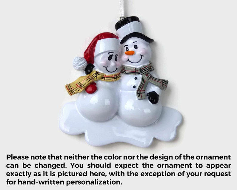May include: A white ceramic Christmas ornament depicting a snowman couple. The female snowman is wearing a red hat and a plaid scarf, while the male snowman is wearing a black top hat and a gray scarf. Both snowmen are smiling and have black eyes and buttons. The ornament is on a white cloud-shaped base.