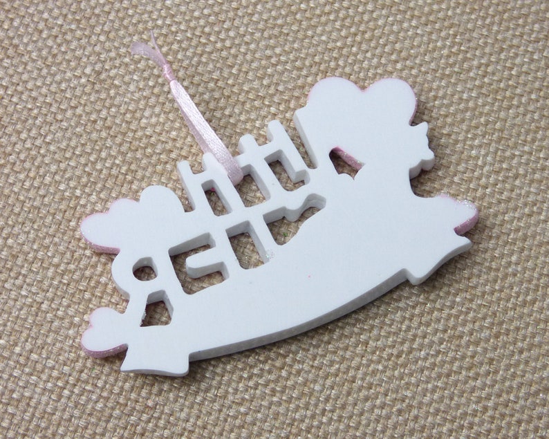May include: White wooden ornament shaped like a rocking horse with pink glitter accents. The ornament has the word "Little" cut out in the center.