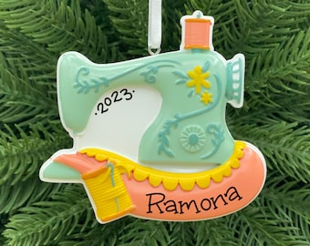 May include: A green and pink sewing machine Christmas ornament with a yellow spool of thread and the name "Ramona" and the year "2023" written on it.