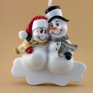May include: A white ceramic Christmas ornament depicting a snowman couple. The male snowman is wearing a black top hat and a gray and red plaid scarf. The female snowman is wearing a red and white Santa hat and a yellow and red plaid scarf. They are both smiling and holding each other.