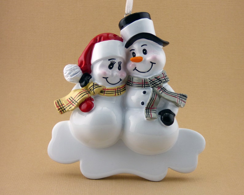 May include: A white ceramic Christmas ornament depicting a snowman couple. The male snowman is wearing a black top hat and a gray and red plaid scarf. The female snowman is wearing a red and white Santa hat and a yellow and red plaid scarf. They are both smiling and holding each other.