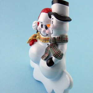 May include: A pair of white ceramic snowman figurines with red, yellow, and black accents. The snowman on the left is wearing a red and white hat and a yellow and black plaid scarf. The snowman on the right is wearing a black top hat and a gray and black plaid scarf.