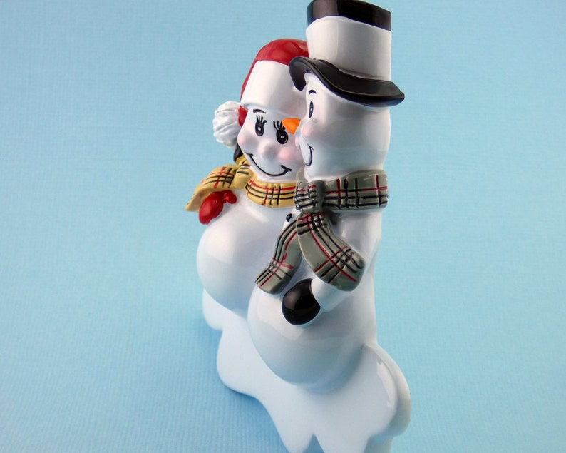 May include: A pair of white ceramic snowman figurines with red, yellow, and black accents. The snowman on the left is wearing a red and white hat and a yellow and black plaid scarf. The snowman on the right is wearing a black top hat and a gray and black plaid scarf.