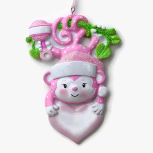 K&ouml;nnte beinhalten: Rosa-wei&szlig;er Weihnachtsbaumschmuck in Form eines Affen, der ein herzf&ouml;rmiges, leeres Schild h&auml;lt. Der Affe tr&auml;gt eine Weihnachtsm&uuml;tze und hat gr&uuml;ne Bl&auml;tter und eine Rassel hinter sich.