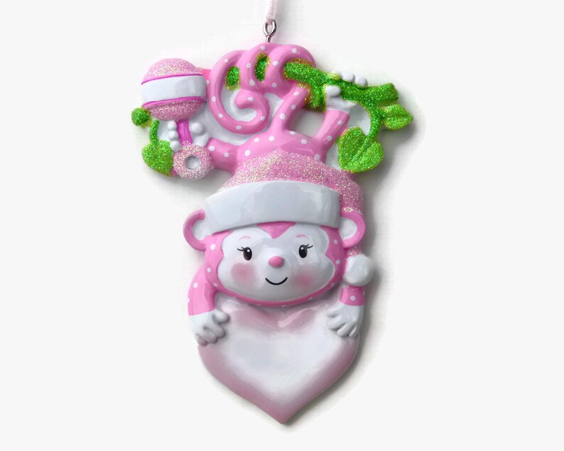 K&ouml;nnte beinhalten: Rosa-wei&szlig;er Weihnachtsbaumschmuck in Form eines Affen, der ein herzf&ouml;rmiges, leeres Schild h&auml;lt. Der Affe tr&auml;gt eine Weihnachtsm&uuml;tze und hat gr&uuml;ne Bl&auml;tter und eine Rassel hinter sich.