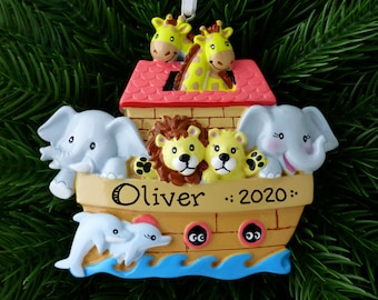 May include: A yellow, brown, and blue ceramic Christmas ornament shaped like Noah's Ark with a red roof. The ark has two elephants, two lions, two giraffes, and two dolphins on it. The name "Oliver" and the year "2020" are written on the front of the ark.