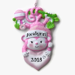 K&ouml;nnte beinhalten: Rosaer Weihnachtsbaumschmuck in Form eines Affen mit einer rosa-wei&szlig;en Rassel, einem gr&uuml;nen Ranken, dem Namen "Avalynn" und dem Jahr "2018" auf einem rosa Herzen.