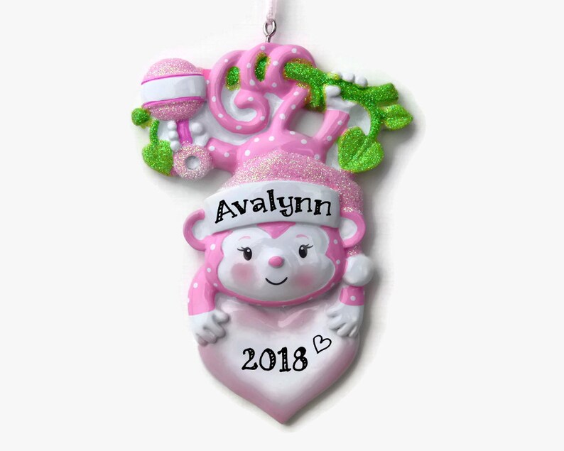 K&ouml;nnte beinhalten: Rosaer Weihnachtsbaumschmuck in Form eines Affen mit einer rosa-wei&szlig;en Rassel, einem gr&uuml;nen Ranken, dem Namen "Avalynn" und dem Jahr "2018" auf einem rosa Herzen.