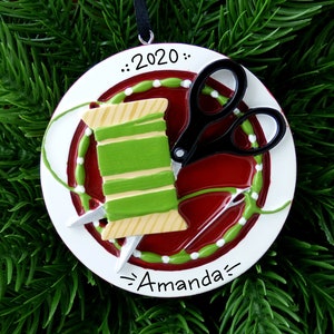 May include: A white round Christmas ornament with a red background and a green and yellow spool of thread, black scissors, and a needle and thread. The ornament has white dots around the edge and the text "...2020..." and "~Amanda~" on it.