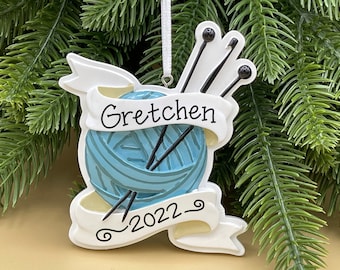 May include: A white ceramic Christmas ornament shaped like a ball of yarn with knitting needles sticking out. The yarn is blue and the ornament has a banner that says "Gretchen 2022".
