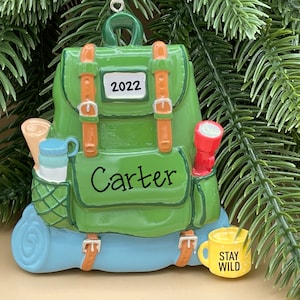 May include: Green backpack Christmas ornament with brown straps, a blue sleeping bag, a red flashlight, a blue water bottle, a brown map, and a yellow mug that says "Stay Wild". The backpack has the name "Carter" and the year "2022" on it.