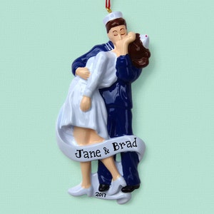 May include: A white and blue Christmas ornament depicting a sailor kissing a nurse. The ornament has a white banner with the text "Jane & Brad" and the year "2017".