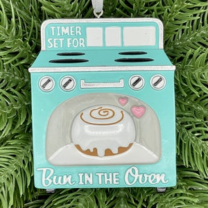 May include: A turquoise blue and silver oven ornament with a cinnamon roll inside. The oven has a timer that says "Timer Set For" and the bottom says "Bun in the Oven".
