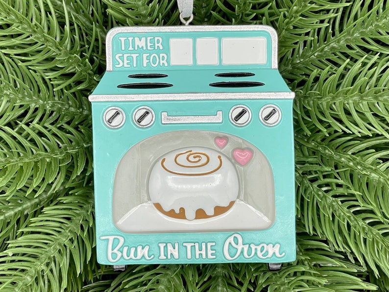 May include: A turquoise blue and silver oven ornament with a cinnamon roll inside. The oven has a timer that says "Timer Set For" and the bottom says "Bun in the Oven".