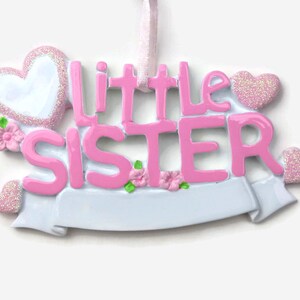 May include: Pink and white ceramic ornament with the words "Little Sister" and a banner. The ornament is decorated with glitter and small pink flowers.