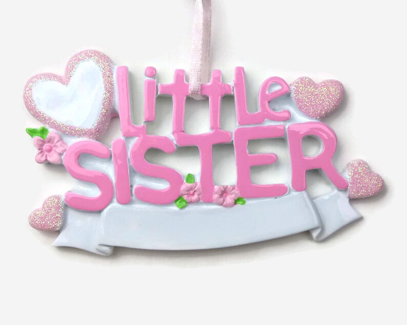 May include: Pink and white ceramic ornament with the words "Little Sister" and a banner. The ornament is decorated with glitter and small pink flowers.