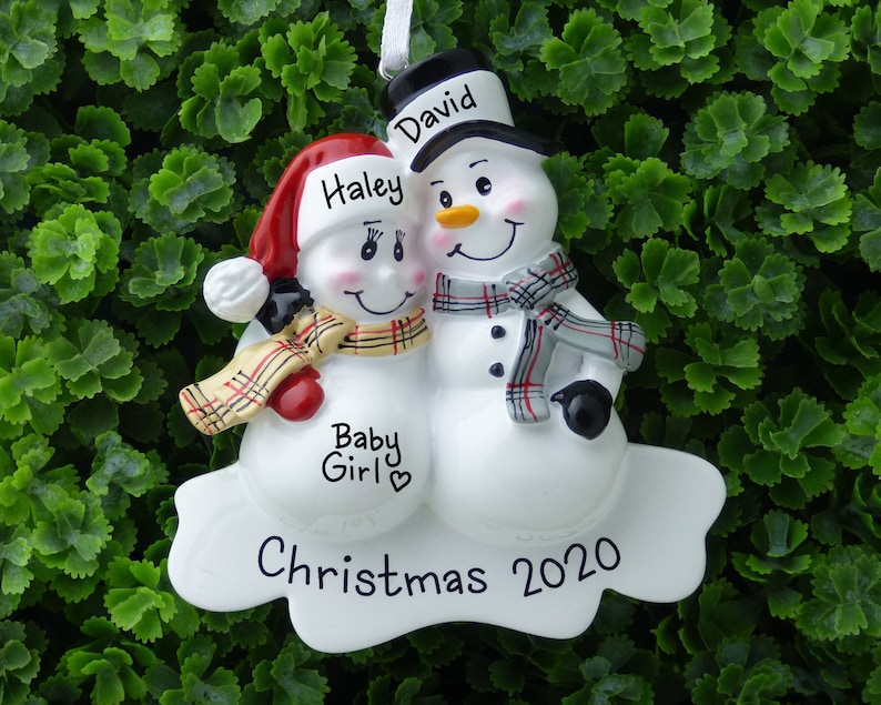 May include: White snowman couple Christmas ornament with red and black hats, plaid scarves, and the names "Haley" and "David" written on them. The ornament also says "Baby Girl" and "Christmas 2020".