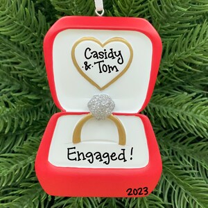 May include: A red and white ornament shaped like an open ring box. The ornament features a gold ring with a silver gem, the words "Cassidy & Tom" inside a heart, and the word "Engaged!" with the year "2023".