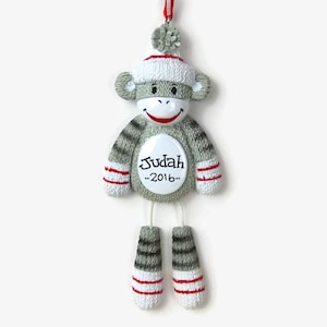 May include: A gray sock monkey Christmas ornament with red and white striped socks, a white hat with a red pom-pom, and a white circle with the name "Judah" and the year "2016" written in black.