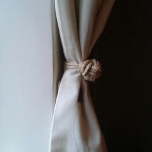 May include: A beige curtain with a natural fiber rope tieback. The rope is tied in a knot with a decorative ball at the end.