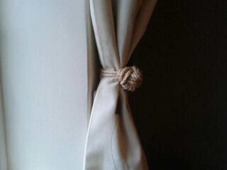 May include: A beige curtain with a natural fiber rope tieback. The rope is tied in a knot with a decorative ball at the end.