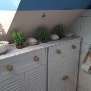 May include: Two white cabinets with gold knobs and slatted doors. The cabinets are decorated with artificial grass plants and seashells.