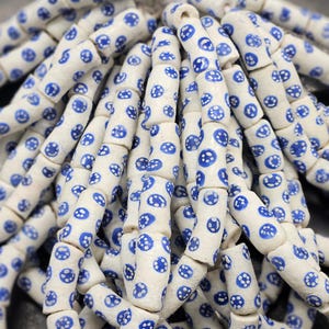 Op de afbeelding: Close-up van witte keramische kralen met een blauw stippenpatroon. De cilindrische kralen zijn aan elkaar geregen, waardoor een textuurontwerp ontstaat. Deze kralen worden waarschijnlijk gebruikt voor het maken van sieraden.