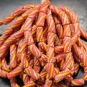 Può includere: Primo piano di una collana di perline colorate. Le perline cilindriche presentano una base rossa con strisce diagonali gialle e piccoli punti bianchi. Le perline sono infilate, creando un aspetto strutturato e vibrante. Ideale per la creazione di gioielli.