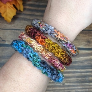 May include: Five crocheted bracelets in a rainbow of colors, including blue, green, purple, orange, and brown. The bracelets are stacked on a person's wrist.
