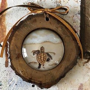Może przedstawiać: Drewniana ozdoba z okrągłym wstawką ze szkła przedstawiającą małego żółwia morskiego na piaszczystej plaży. Ozdoba jest związana brązową wstążką.