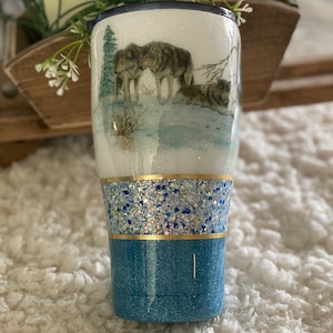 Puede incluir: Un vaso de glitter blanco y azul con una escena nevada con dos lobos. El vaso tiene una banda de glitter dorado y una base de glitter azul.